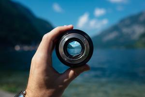 Individual holding a camera lens with mountains and lake in the distance